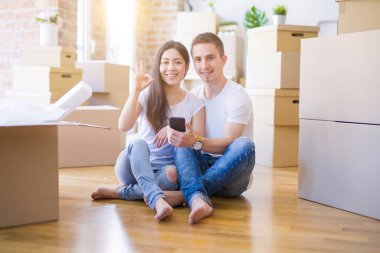 Beautiful couple sitting on the floor using smartphone at new home around cardboard boxes doing ok sign with fingers, excellent symbol