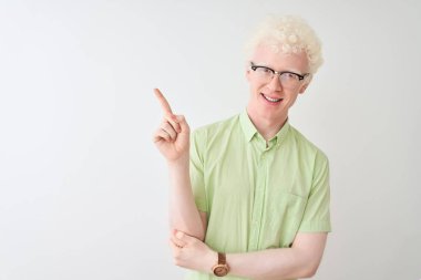Yeşil gömlekli ve gözlüklü genç albino bir adam izole edilmiş beyaz arka planda, yüzünde kocaman bir gülümseme, eli ve parmağı kameraya bakıyor..
