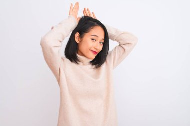 Young chinese woman wearing turtleneck sweater standing over isolated white background Doing bunny ears gesture with hands palms looking cynical and skeptical. Easter rabbit concept.