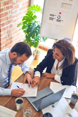 Two middle age business workers smiling happy and confident. Working together with smile on face using laptop at the office clipart