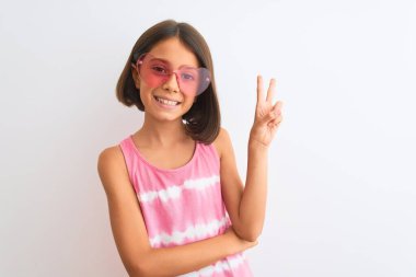 İzole edilmiş beyaz arka plan üzerinde pembe tişört ve güneş gözlüğü takan güzel bir kız çocuğu. Kameraya gülümseyen ve zafer işareti yapan bir gülücük. İki numara..