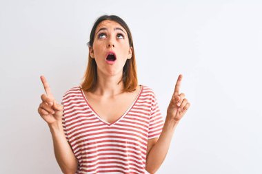 Beautiful redhead woman wearing casual striped red t-shirt over isolated background amazed and surprised looking up and pointing with fingers and raised arms.