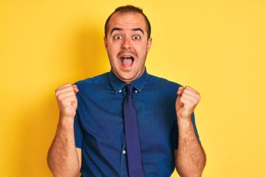 Young businessman wearing denim casual shirt and tie over isolated yellow background celebrating surprised and amazed for success with arms raised and open eyes. Winner concept.
