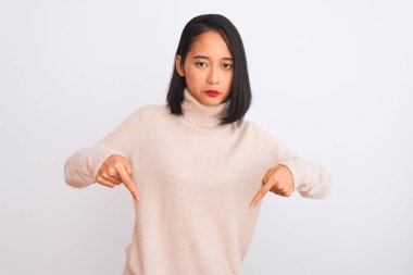 Young chinese woman wearing turtleneck sweater standing over isolated white background Pointing down looking sad and upset, indicating direction with fingers, unhappy and depressed.