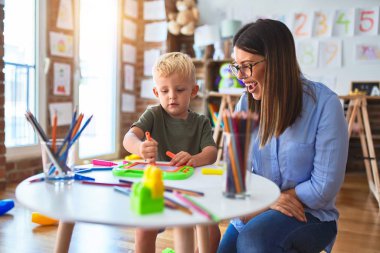 Genç beyaz çocuk öğretmeniyle oyun okulunda oynuyor. Anne ve oğlu oyun odasında renkli kalemlerle beraberlik çiziyor.