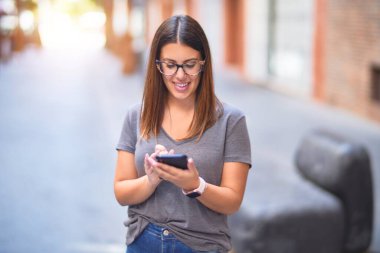 Mutlu ve kendinden emin gülümseyen genç ve güzel bir kadın. Yüzünde gülümsemeyle şehrin caddesinde akıllı telefon kullanarak dikiliyorsun.