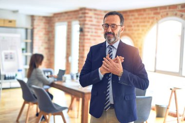 Middle age handsome businessman wearing glasses standing at the office smiling with hands on chest with closed eyes and grateful gesture on face. Health concept. clipart