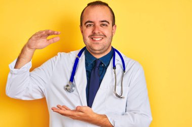 Young doctor man wearing coat and stethoscope standing over isolated yellow background gesturing with hands showing big and large size sign, measure symbol. Smiling looking at the camera. Measuring concept.