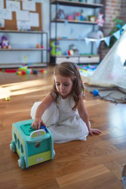 Anaokulunda bir sürü oyuncağın etrafında arabayla oynayan sevimli sarışın çocuk.