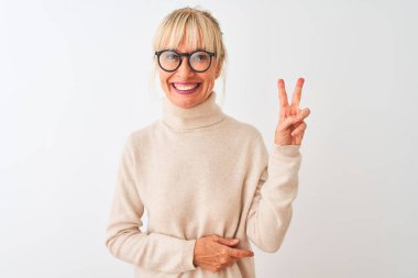 Balıkçı yaka kazak ve gözlük takan orta yaşlı bir kadın izole edilmiş beyaz arka planda gülümsüyor ve kameraya gülümsüyor zafer işareti yapıyor. İki numara..