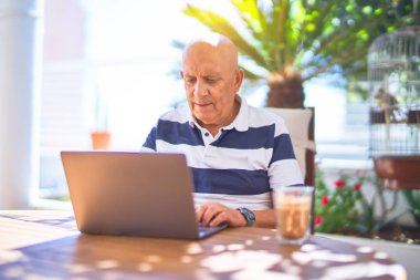 Kıdemli yakışıklı adam mutlu ve kendinden emin gülümsüyor. Terasta dizüstü bilgisayar kullanarak oturmak