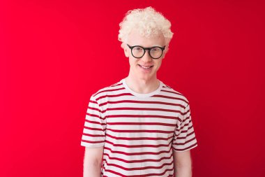 Genç albino sarışın adam çizgili tişört ve gözlüklerle izole edilmiş kırmızı arka planda mutlu ve havalı bir gülümseme takınıyor. Şanslı kişi..