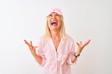 İzole edilmiş beyaz arka plan üzerinde uyku maskesi ve pijama giyen orta yaşlı bir kadın çılgın ve sinirli bir şekilde bağırıyor ve agresif bir ifade ve kolla bağırıyor. Hayal kırıklığı konsepti.
