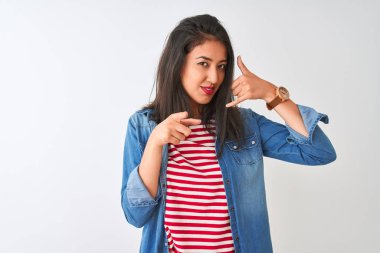 Young chinese woman wearing striped t-shirt and denim shirt over isolated white background smiling doing talking on the telephone gesture and pointing to you. Call me.