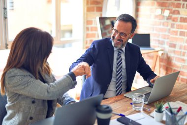 Two middle age business workers smiling happy and confident. Working together with smile on face giving fist bump at the office clipart