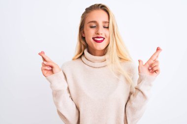 Balıkçı yaka kazak giyen genç ve güzel bir kadın izole edilmiş beyaz arka plan üzerinde durup umut ve gözler kapalı gülümseyerek el sallıyor. Şans ve batıl inanç..