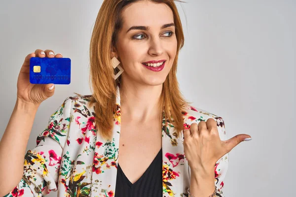 Redhead caucasian business woman holding credit card over isolated background pointing and showing with thumb up to the side with happy face smiling