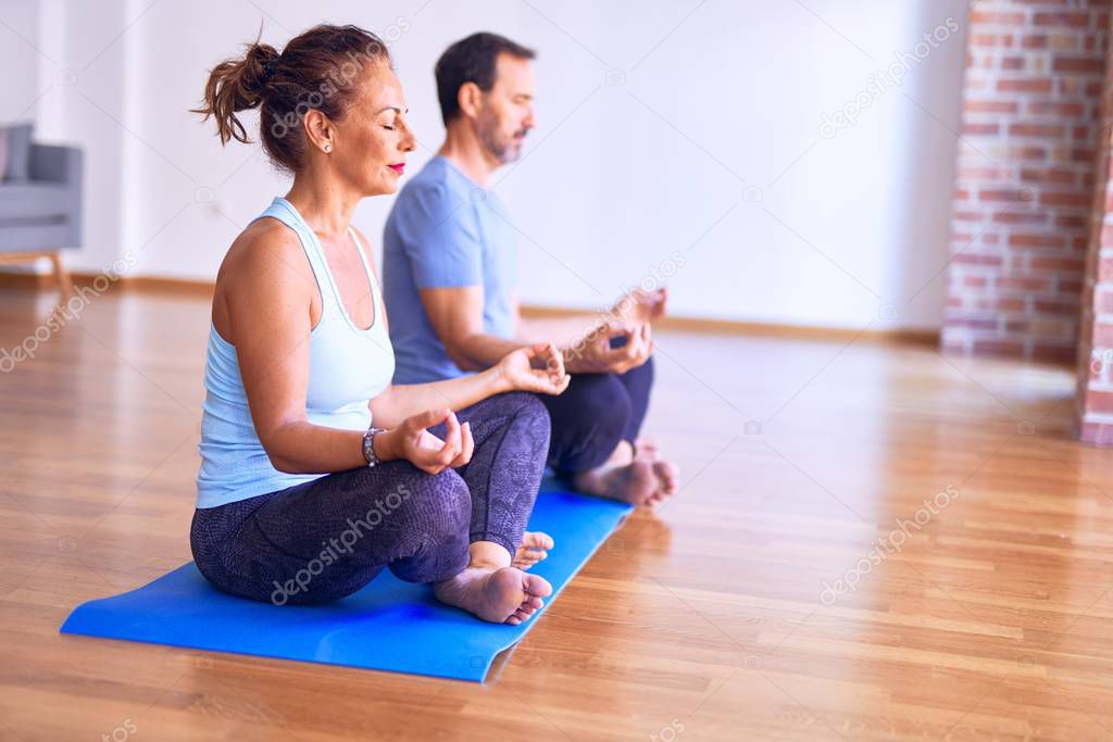 Middle age beautiful sporty couple sitting on mat practicing yoga doing lotus pose at gym