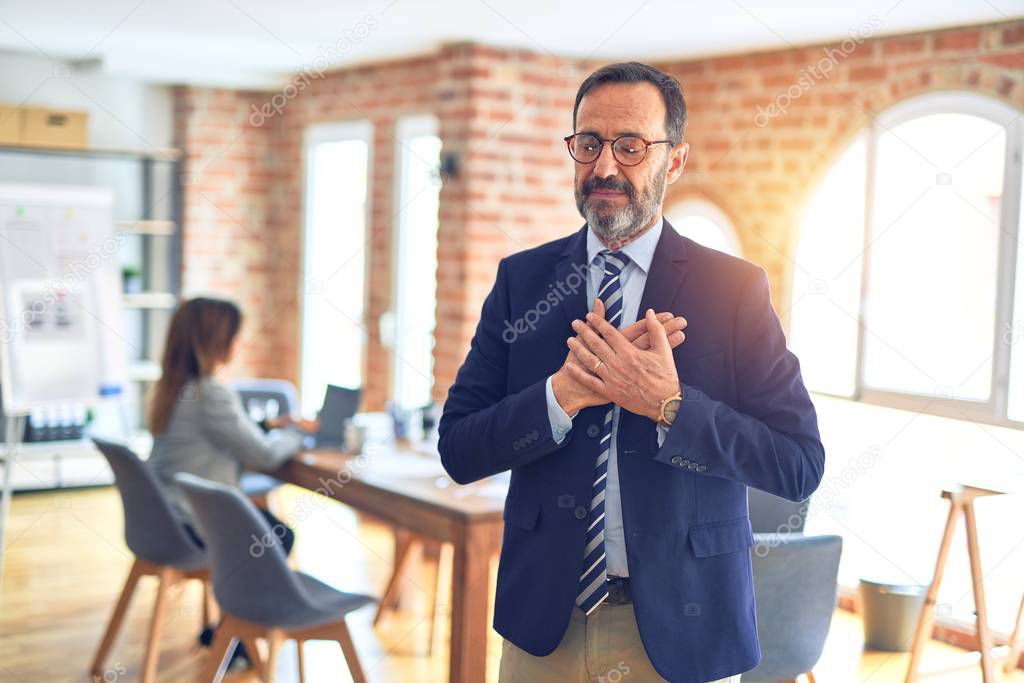 Middle age handsome businessman wearing glasses standing at the office smiling with hands on chest with closed eyes and grateful gesture on face. Health concept.