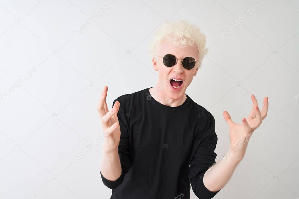 Young albino man wearing black t-shirt and sunglasess standing over isolated white background crazy and mad shouting and yelling with aggressive expression and arms raised. Frustration concept.