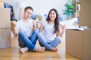 Genç ve güzel bir çift, yeni evlerinde, mukavva kutuların etrafında oturmuş, mutlu ve heyecanlı bir şekilde kollarını kaldırıp, gülümseyerek ve başarı çığlıkları atarak kazanan jestler yapıyorlar. Kutlama konsepti.