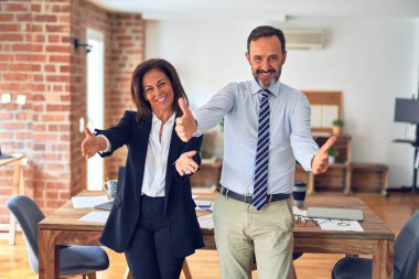 Two middle age business workers standing working together in a meeting at the office looking at the camera smiling with open arms for hug. Cheerful expression embracing happiness. clipart