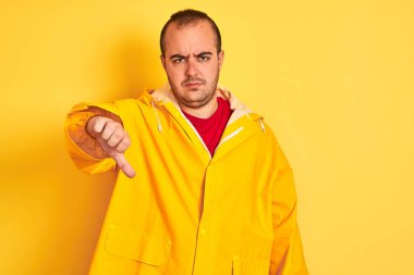 Young man wearing rain coat standing over isolated yellow background looking unhappy and angry showing rejection and negative with thumbs down gesture. Bad expression.
