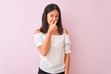 Beautiful chinese woman wearing white t-shirt standing over isolated pink background smelling something stinky and disgusting, intolerable smell, holding breath with fingers on nose. Bad smells concept.