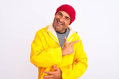 Orta yaşlı bir adam, yağmurluk ve yünlü şapka giyer, izole edilmiş beyaz arka planda neşeli durur, yüzünde mutlu ve doğal bir ifadeyle, elleri ve parmaklarıyla yüzünü gösteren bir gülümseme vardır.