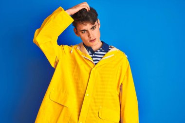 Teenager fisherman boy wearing yellow raincoat over isolated background confuse and wonder about question. Uncertain with doubt, thinking with hand on head. Pensive concept.