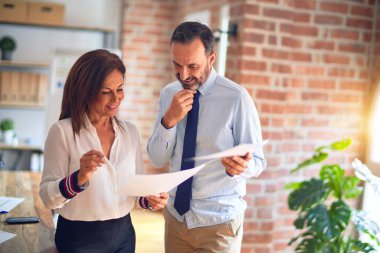 Two middle age business workers smiling happy and confident. Standing with smile on face working together holding documents at the office clipart