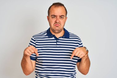 Young man wearing casual striped polo standing over isolated white background Pointing down looking sad and upset, indicating direction with fingers, unhappy and depressed.