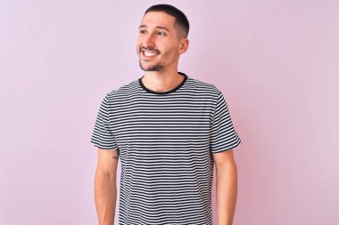 Çizgili tişört giyen genç yakışıklı adam pembe izole edilmiş arka plan üzerinde duruyor yüzünde gülümseme, doğal bir ifade ile yan yana bakıyor. Kendinden emin gülen.