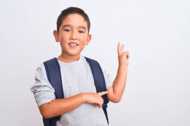 Güzel öğrenci çocuk sırt çantası takıp izole edilmiş beyaz arka planda dikiliyor. Kameraya bakıp gülümsüyor ve zafer işareti yapıyor. İki numara..