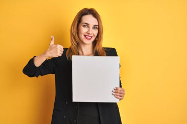 Young redhead businesswoman with clear eyes holding banner over isolated yellow background happy with big smile doing ok sign, thumb up with fingers, excellent sign