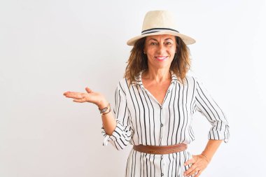 Orta yaşlı iş kadını çizgili elbise ve şapkalı, izole edilmiş beyaz arka planda neşeli bir şekilde gülümseyen ve avuç içi kameraya bakan..