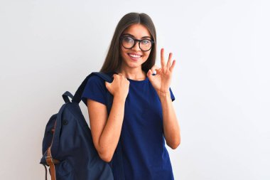 Sırt çantası ve gözlük takan genç ve güzel bir bayan izole edilmiş beyaz arka plan üzerinde tamam işareti yapıyor, mükemmel bir sembol.