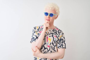 Renkli tişört ve güneş gözlüğü takan genç albino bir adam izole edilmiş kırmızı arka planda, elleri çenesinde soru sorarken, düşünceli bir ifade. Düşünceli bir yüzle gülümsüyor. Şüphe kavramı.