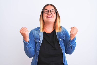 Kotlu tişört ve gözlük takan genç ve güzel bir kadın izole edilmiş beyaz arka planda şaşırmış ve şaşırmış bir şekilde kollarını kaldırıp, gözlerini açarak başarıyı kutluyor. Kazanan konsept.