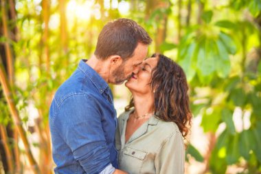 Middle age beautiful couple smiling happy and confident at town park. Standing with smile on face hugging and kissing clipart