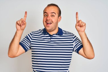 Young man wearing casual striped polo standing over isolated white background smiling amazed and surprised and pointing up with fingers and raised arms.