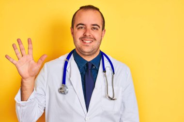 Young doctor man wearing coat and stethoscope standing over isolated yellow background showing and pointing up with fingers number five while smiling confident and happy.