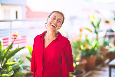 Orta yaşlı güzel bir kadın mutlu ve kendinden emin gülümsüyor terasta yüzünde bir gülümsemeyle duruyor.