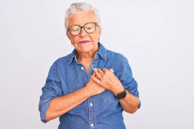 Yaşlı, gri saçlı, kot gömlek ve gözlüklü bir kadın izole edilmiş beyaz arka plan üzerinde elleri göğsünde, gözleri kapalı ve yüzünde minnet dolu bir jest ile gülümsüyor. Sağlık kavramı.