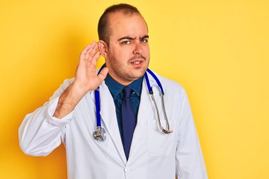 Young doctor man wearing coat and stethoscope standing over isolated yellow background smiling with hand over ear listening an hearing to rumor or gossip. Deafness concept.