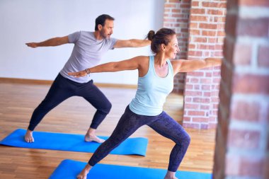Orta yaşlı güzel sporcu çift mutlu gülümsüyor. Yüzünde gülümsemeyle ayakta durup, spor salonunda savaşçı pozu vererek yoga yapıyor.