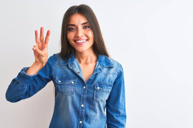 İzole edilmiş beyaz arka plan üzerinde duran sıradan bir kot gömlek giyen genç ve güzel bir kadın üç numaralı parmaklarıyla işaret ederken kendinden emin ve mutlu gülümsüyor..