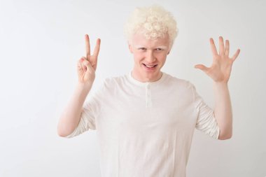 Genç albino tişörtlü sarışın adam izole edilmiş beyaz arka plan üzerinde duruyor ve yedinci parmaklarını gösterirken kendinden emin ve mutlu bir şekilde gülümsüyor..