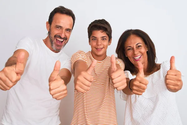 Family of three, mother, father and son standing over white isolated background approving doing positive gesture with hand, thumbs up smiling and happy for success. Winner gesture.
