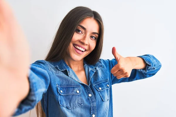 Beautiful woman wearing denim shirt make selfie by camera over isolated white background approving doing positive gesture with hand, thumbs up smiling and happy for success. Winner gesture.